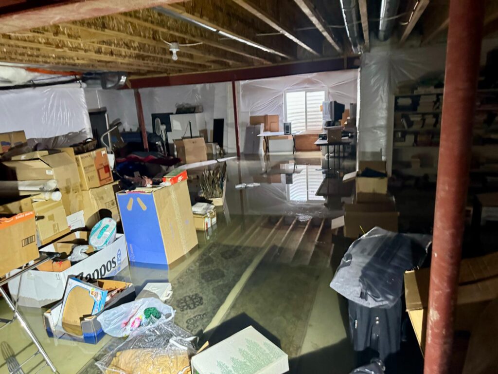 Contaminated flood water covering basement floor with damaged storage boxes, household items, and visible flooding risks in a Denver home.