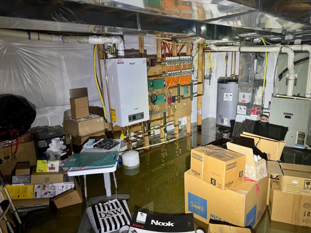 Flooded basement utility room with submerged water heater and electrical systems, surrounded by boxes and household items, highlighting the risks of basement flooding in Denver.