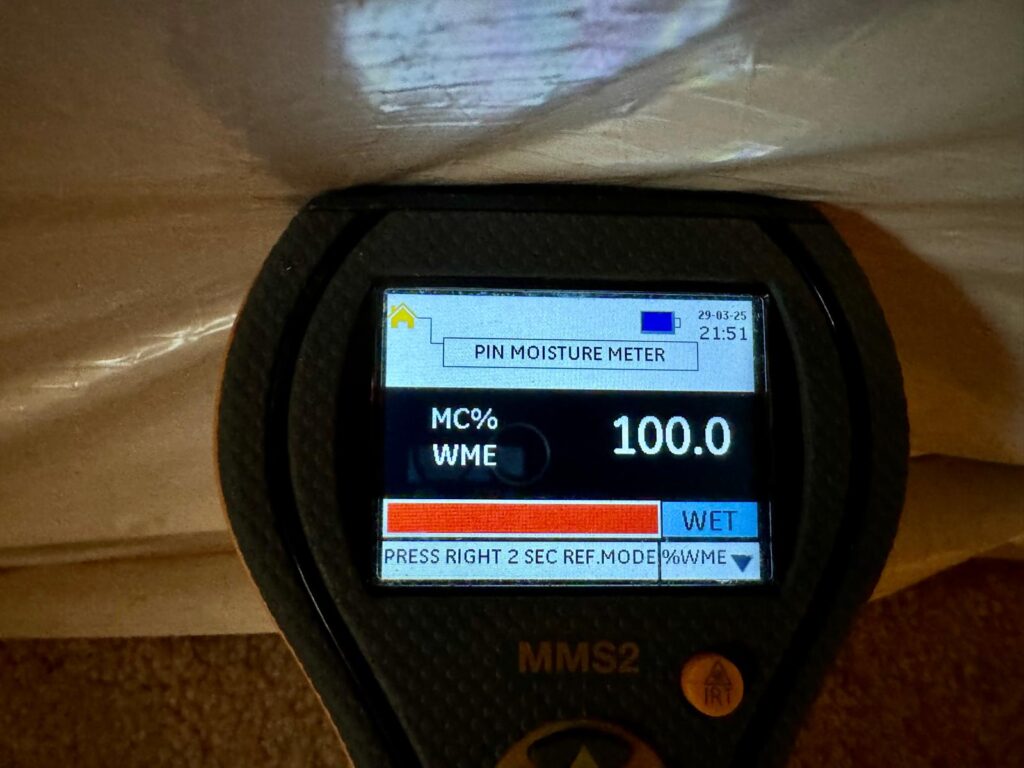 Moisture meter displaying 100% saturation reading, indicating wet building materials after basement flooding.
